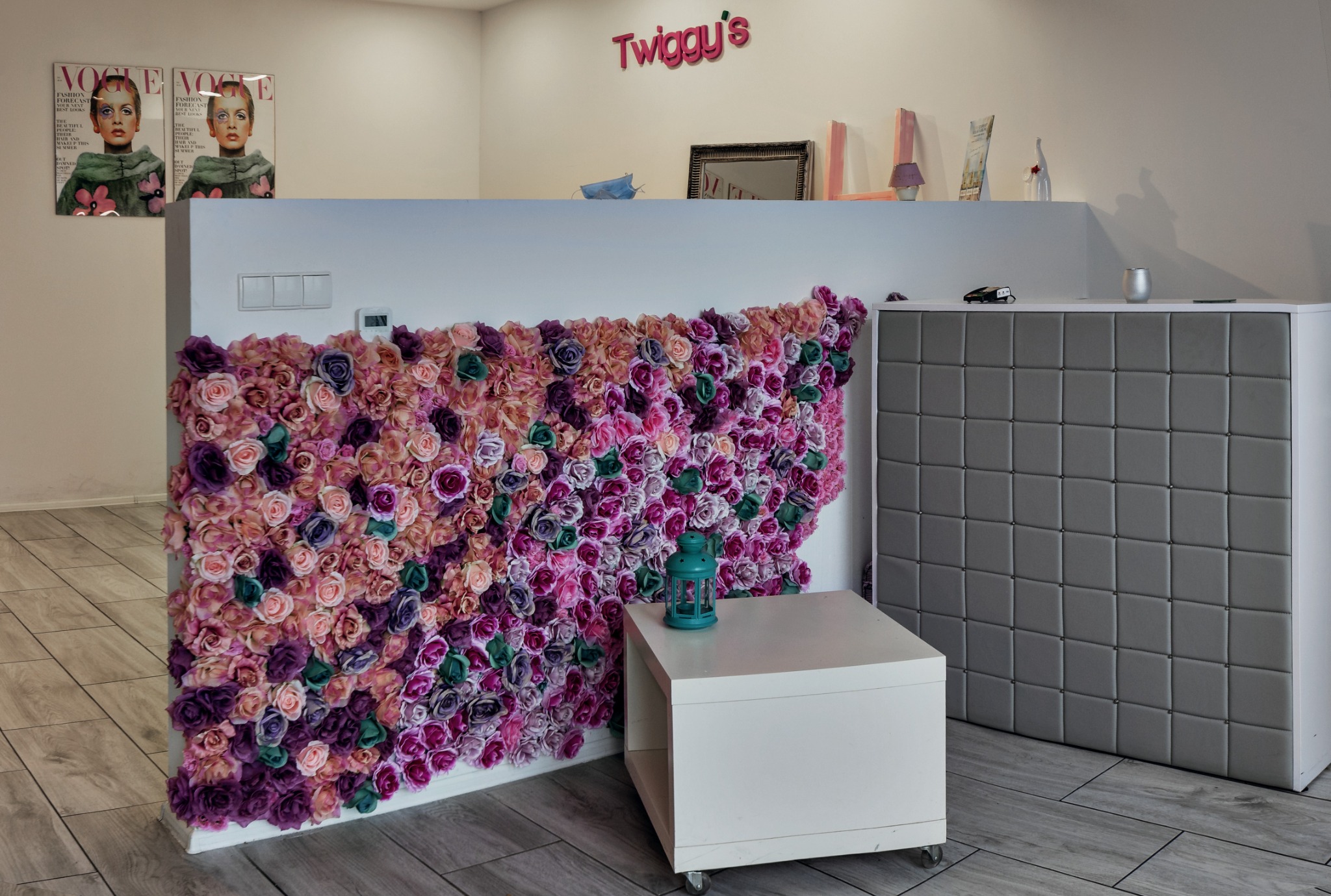 Recepcja salonu z dekoracją z kwiatów w odcieniach fioletu i różu, na ścianie plakaty z Twiggy i logo salonu.