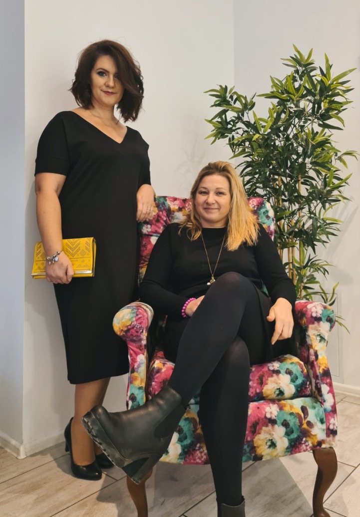 Dwie kobiety w czarnych sukienkach pozują w salonie: jedna stoi trzymając żółtą kopertówkę, druga siedzi w fotelu w kwiaty, prezentując stylizację.