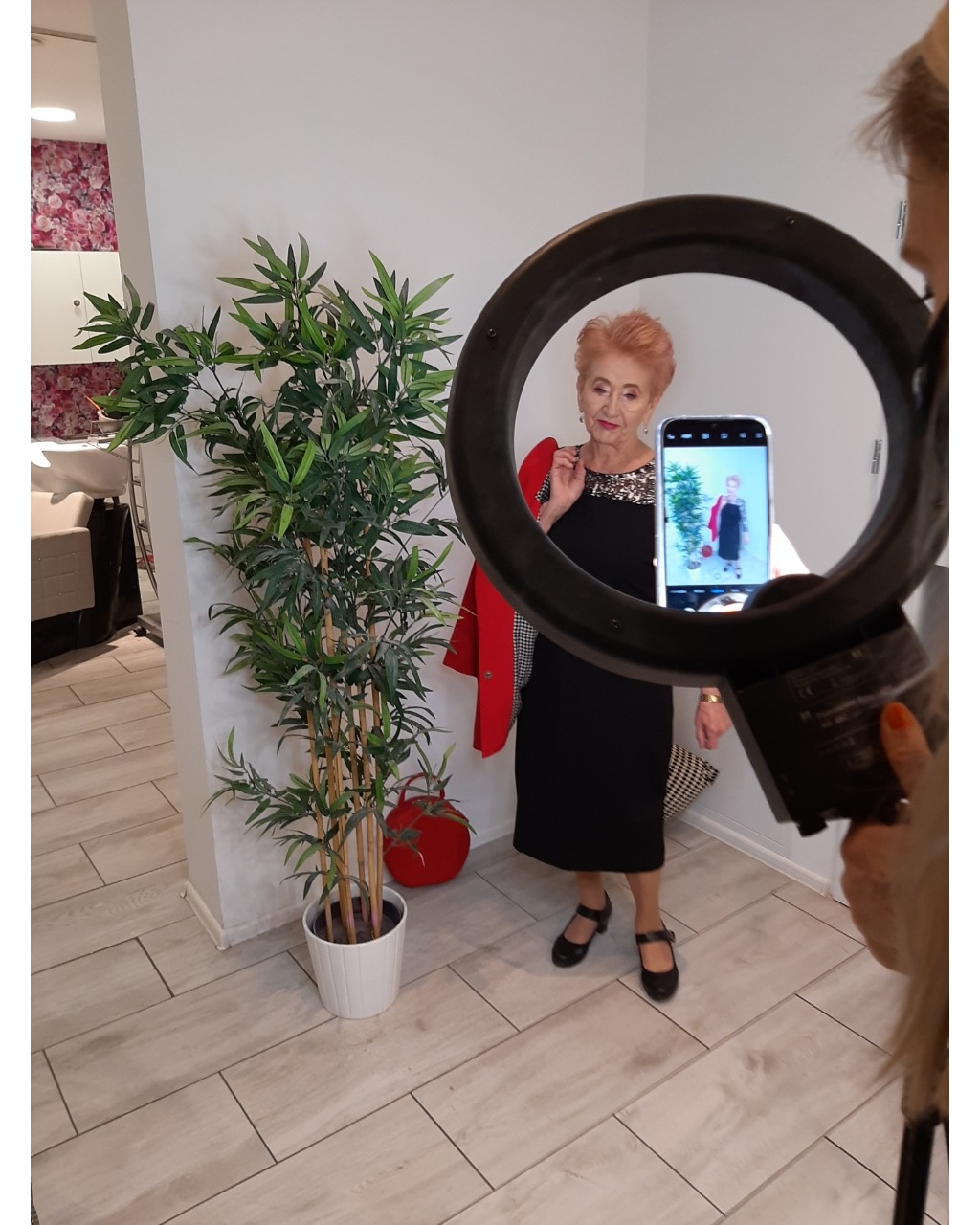 Kobieta w czarnej sukience pozuje do zdjęcia robionego telefonem przez ring light w salonie z rośliną doniczkową i elementami wystroju.