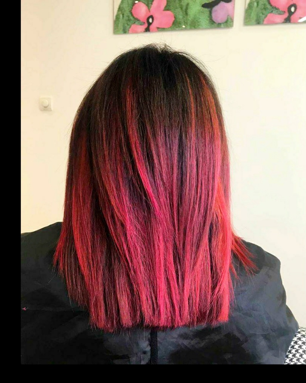 Tył głowy z prostymi, ciemnymi włosami z jaskrawo czerwonymi pasemkami, widoczny czarny fartuch ochronny.