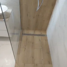 Kabina prysznicowa typu walk-in z odpływem liniowym, wykończona płytkami imitującymi drewno i marmur, z widoczną baterią prysznicową.