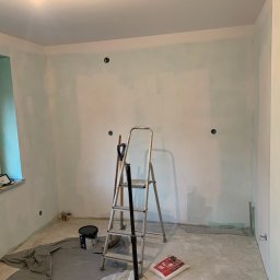 Properbud Sp. z o.o. - Pokój w trakcie przygotowania do malowania: ściany pokryte warstwą podkładową, drabina aluminiowa, wiadro z farbą i worek gładzi na podłodze zabezpieczonej folią malarską.