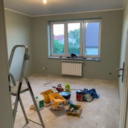 Properbud Sp. z o.o. - Pokój w trakcie malowania na pastelowy odcień zieleni, widoczne drabina, pędzle, wałki i otwarte puszki z farbą na podłodze, okno z widokiem na zabudowania.