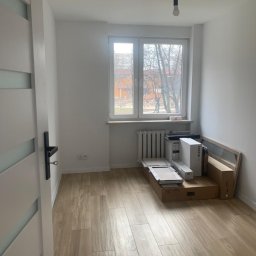 Properbud Sp. z o.o. - Pusty pokój po świeżym malowaniu ścian, z ułożoną podłogą z paneli imitujących drewno, kartony z panelami pod oknem i wisząca żarówka.