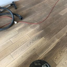 Properbud Sp. z o.o. - Szlifowanie drewnianej podłogi parkietowej maszyną z odkurzaczem, widoczny kabel zasilający i wąż odkurzacza.