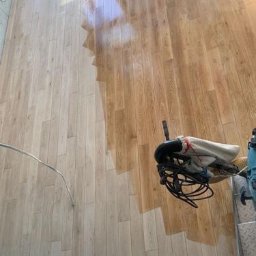 Properbud Sp. z o.o. - Polerowanie drewnianej podłogi parkietowej maszyną, widoczny fragment wypolerowanej, błyszczącej powierzchni, w tle okno i ściana w jasnych barwach.