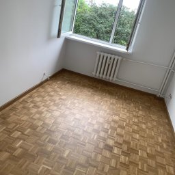 Properbud Sp. z o.o. - Jasny pokój z parkietem w jodełkę, białymi ścianami i grzejnikiem pod oknem z widokiem na drzewa.