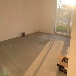 Properbud Sp. z o.o. - Pokój w trakcie przygotowań do malowania: surowe ściany, betonowa podłoga, puszka z farbą przy ścianie, karton z materiałami pod oknem, silne promienie słoneczne wpadające przez okno.