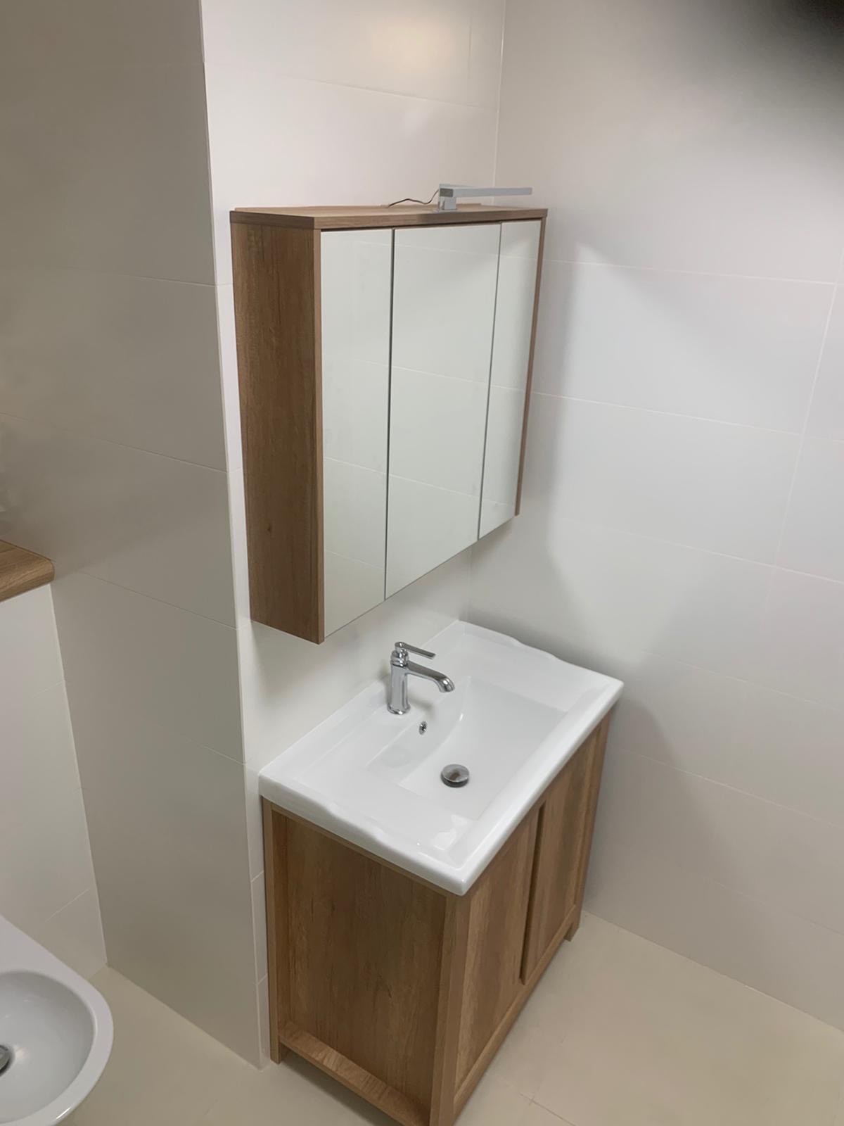 Nowoczesna, jasna łazienka z umywalką w szafce z drewna i szafką lustrzaną z oświetleniem nad nią, wykończona białymi płytkami.