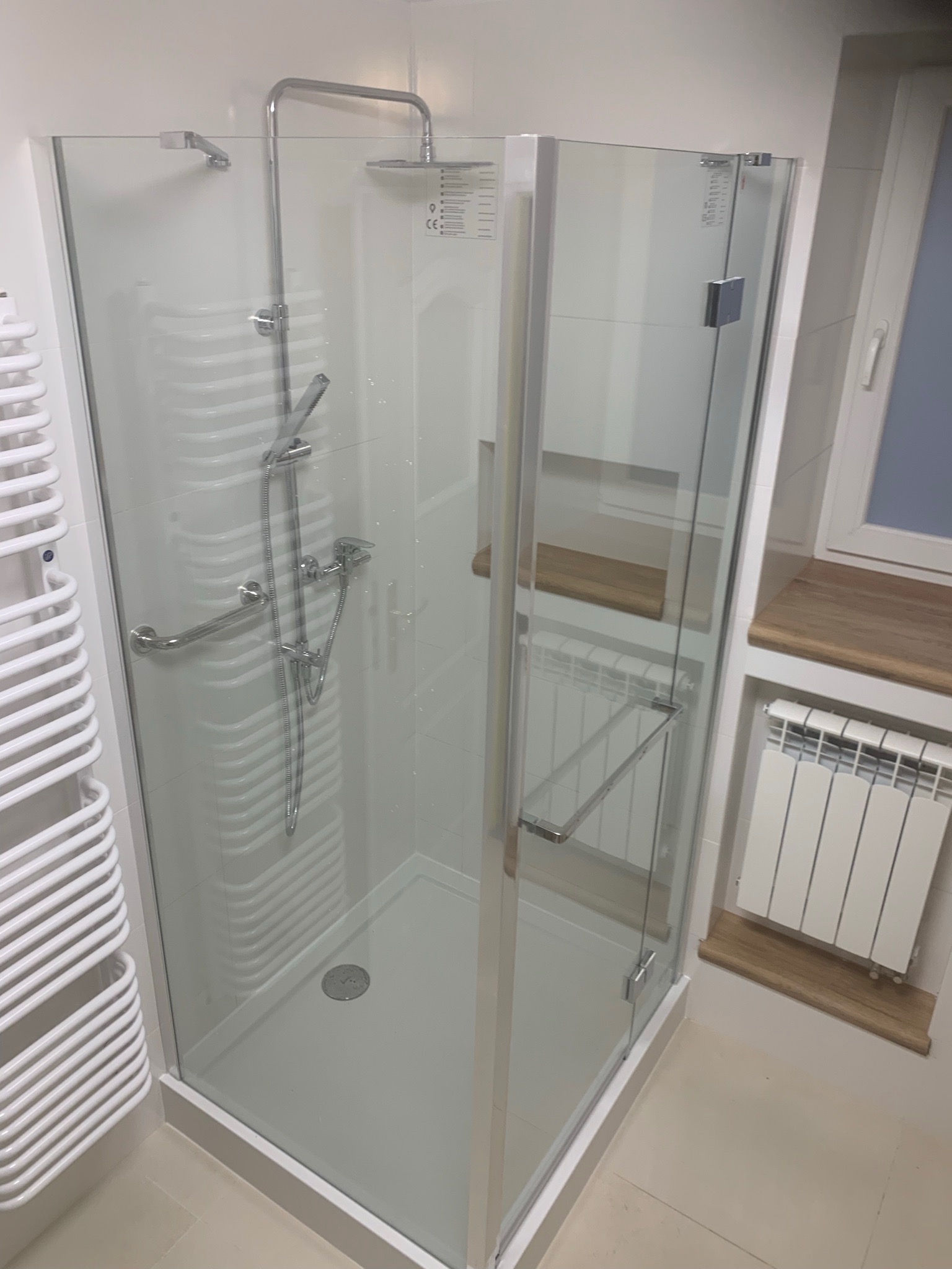 Nowoczesna kabina prysznicowa z chromowaną armaturą, uchwytem dla niepełnosprawnych i grzejnikiem drabinkowym w tle, w jasnej łazience.
