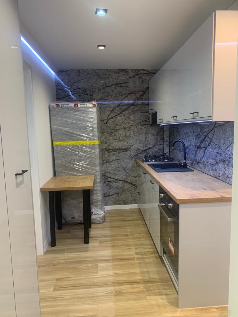 Wąska kuchnia w trakcie wykończenia: białe szafki górne i dolne, drewniany blat, zlewozmywak, płyta grzewcza, piekarnik, marmurowa ściana, podłoga z paneli, stół z czarnymi nogami, lodówka w folii...