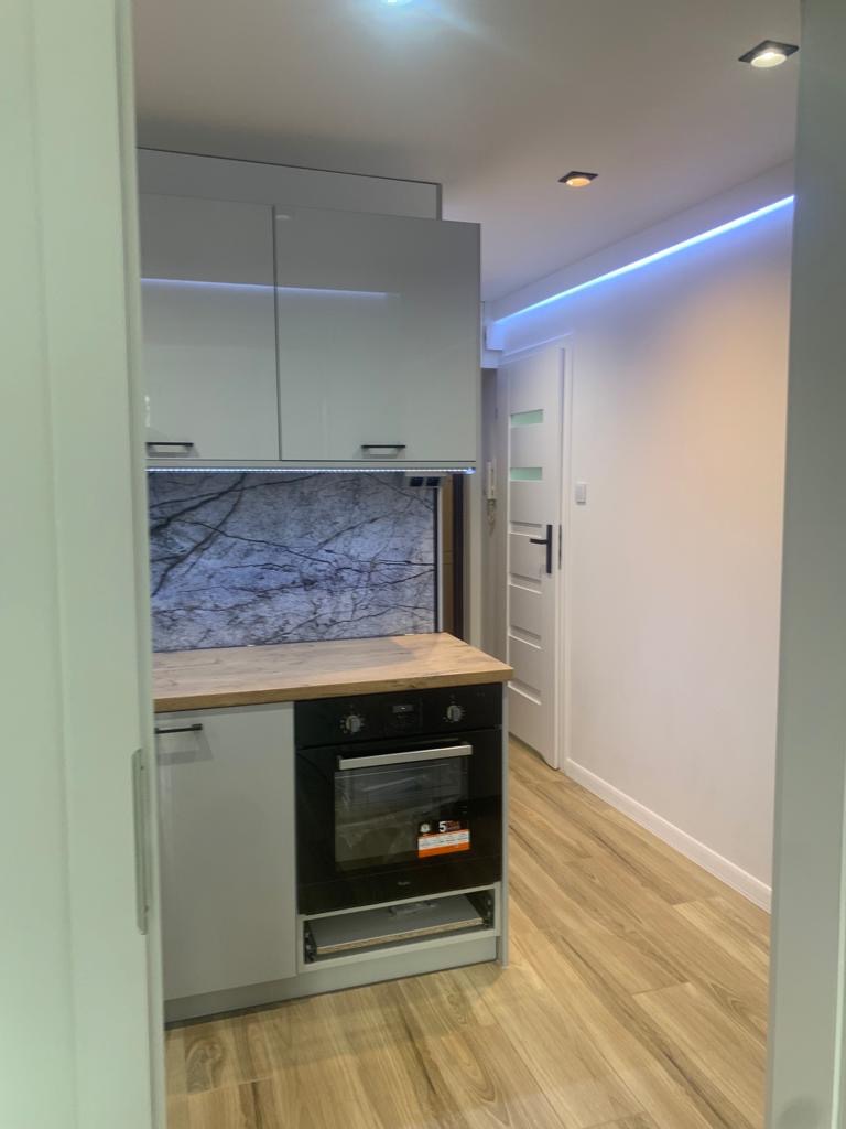 Nowoczesna kuchnia z białymi szafkami, drewnianym blatem i marmurowym panelem nad blatem, oświetlona ledami, widoczna z perspektywy wejścia do pomieszczenia.