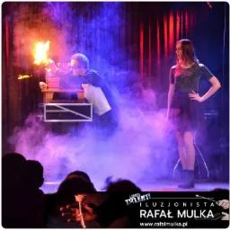 Magik w trakcie pokazu z ogniem w dłoniach, asystentka stoi obok w dymie i niebieskim oświetleniu scenicznym, logo iluzjonisty w dolnej części kadru.