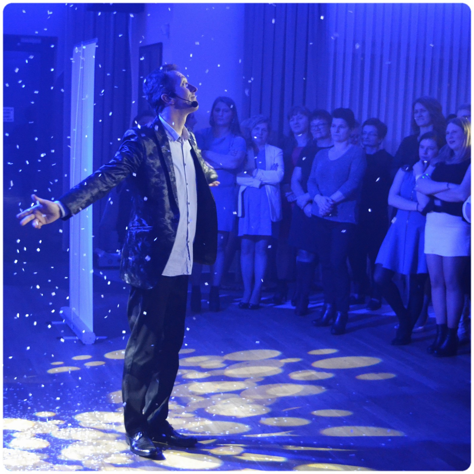 Mężczyzna w marynarce i słuchawkach mikrofonowych, z rozłożonymi rękami, na scenie w niebieskim oświetleniu, z publicznością w tle i opadającymi konfetti.