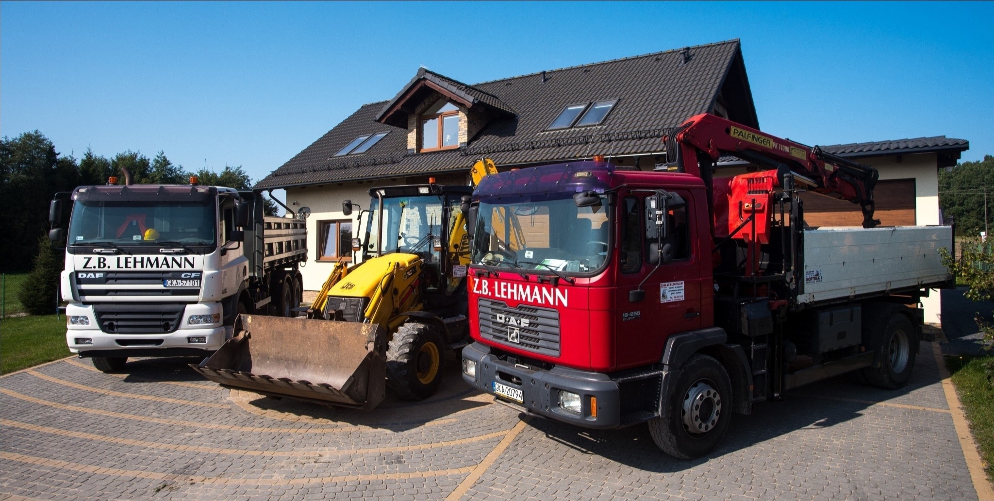 Flota firmy Z.B. Lehmann: biała ciężarówka DAF, żółta koparko-ładowarka JCB i czerwona ciężarówka MAN z dźwigiem Palfinger, zaparkowane na wybrukowanym podjeździe przed domem z ciemną dachówką.