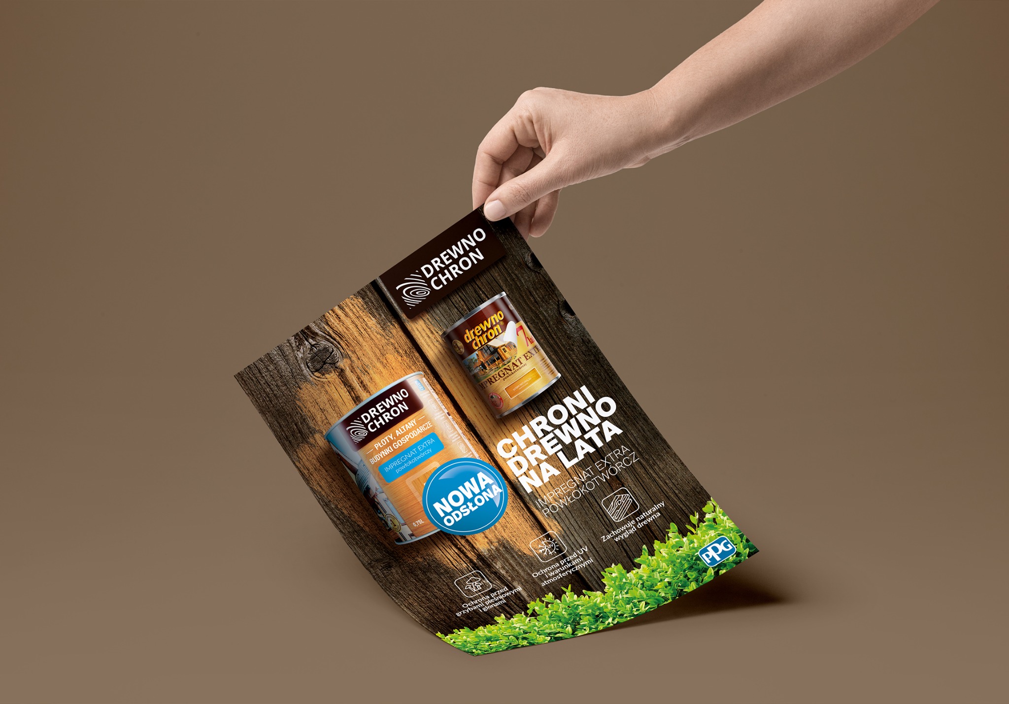 Dłoń trzyma ulotkę reklamową impregnatów do drewna marki Drewnochron, prezentującą puszkę produktu na tle deski i hasło 'Chroni drewno na lata'.