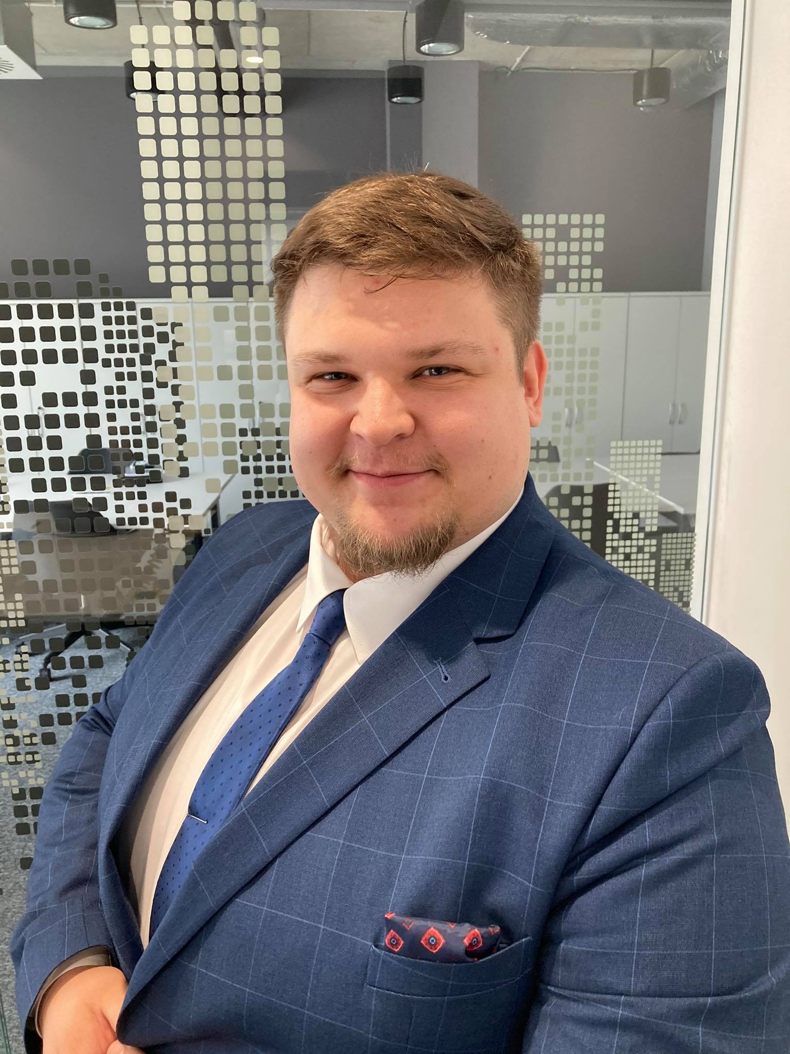 Uśmiechnięty mężczyzna w niebieskim garniturze w kratę, z wzorzystą poszetką w kieszonce, stojący na tle biurowej ściany z geometrycznym wzorem.