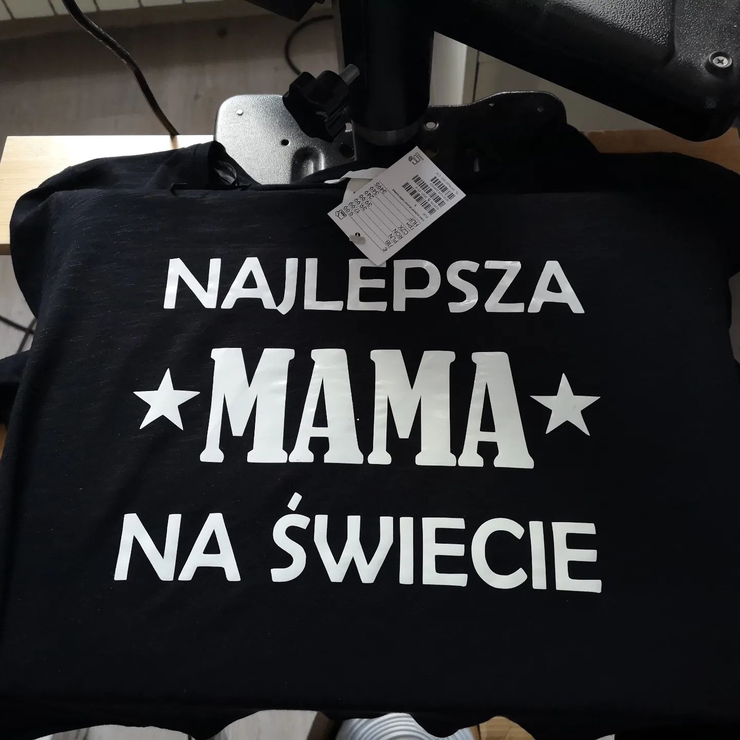 Czarna koszulka z białym napisem 'NAJLEPSZA *MAMA* NA ŚWIECIE' leżąca pod prasą termotransferową, widoczna metka.