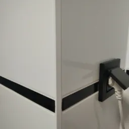 Białe, błyszczące płytki z czarnym pasem dekoracyjnym, narożnik i czarne gniazdko z wtyczką. Minimalistyczne wykończenie ściany w łazience lub kuchni.
