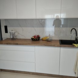 Michał Szymanski - Biała, nowoczesna kuchnia z drewnianym blatem i marmurowym panelem. Na blacie owoce, czajnik i czarna bateria. Widoczny refleks osoby w górnych szafkach.