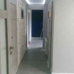 Michał Szymanski - Wykończony korytarz w odcieniach szarości z oświetleniem LED na suficie i imitacją cegły na ścianie. Widoczne drzwi i płytki podłogowe.