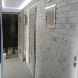 Michał Szymanski - Korytarz po remoncie: biała cegła na ścianie, szare płytki na podłodze, dwoje drzwi wewnętrznych i oświetlenie sufitowe LED. Widoczne puszki elektryczne.