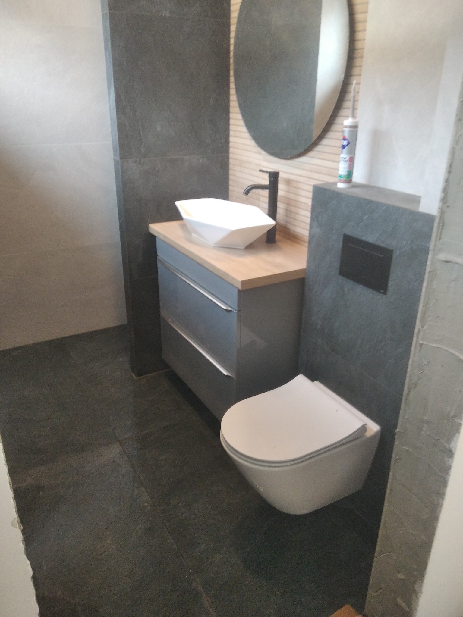 Nowoczesna łazienka z szarą armaturą i podwieszaną toaletą. Umywalka nablatowa o geometrycznym kształcie. Zastosowano płytki w odcieniach szarości.