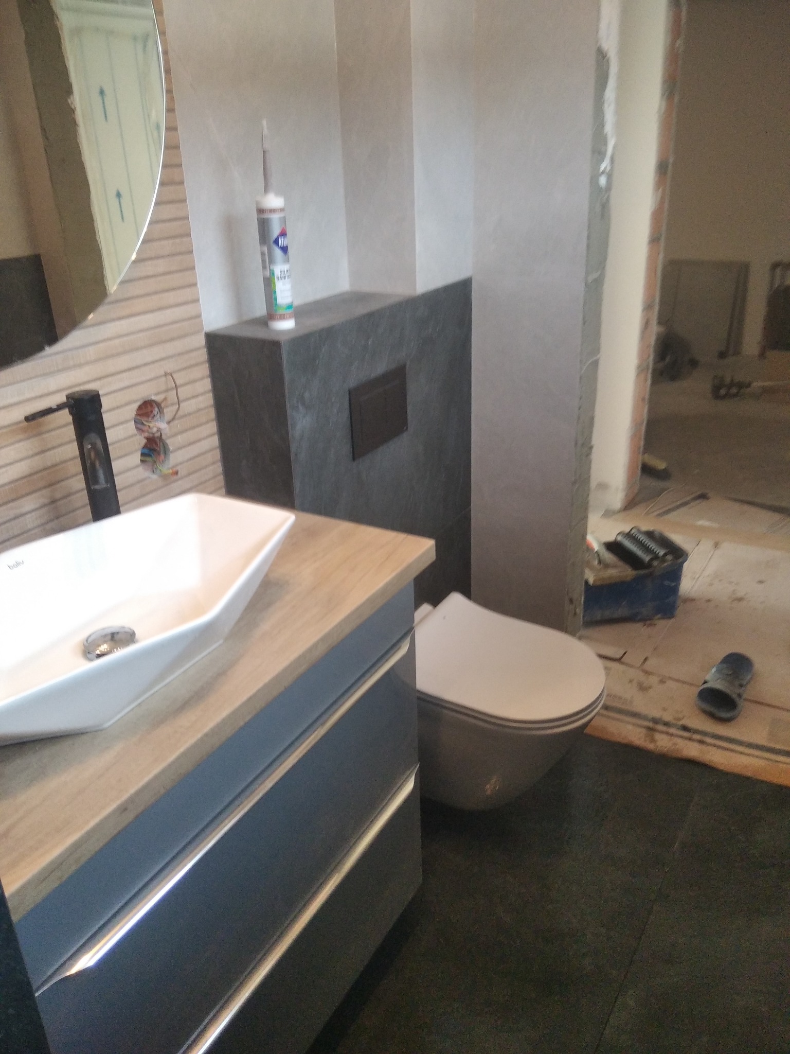 Nowoczesna łazienka w trakcie remontu: biała umywalka nablatowa, szare płytki, widoczne niedokończone instalacje elektryczne i narzędzia. W tle surowa przestrzeń.