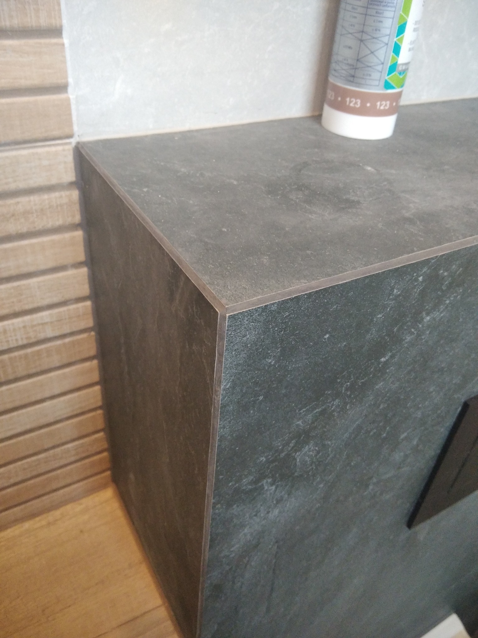 Szara, kamienna zabudowa z precyzyjnie dociętymi płytkami, widoczna tuba silikonu i fragment drewnopodobnej ściany. Estetyczne wykończenie, detale i tekstura materiałów.