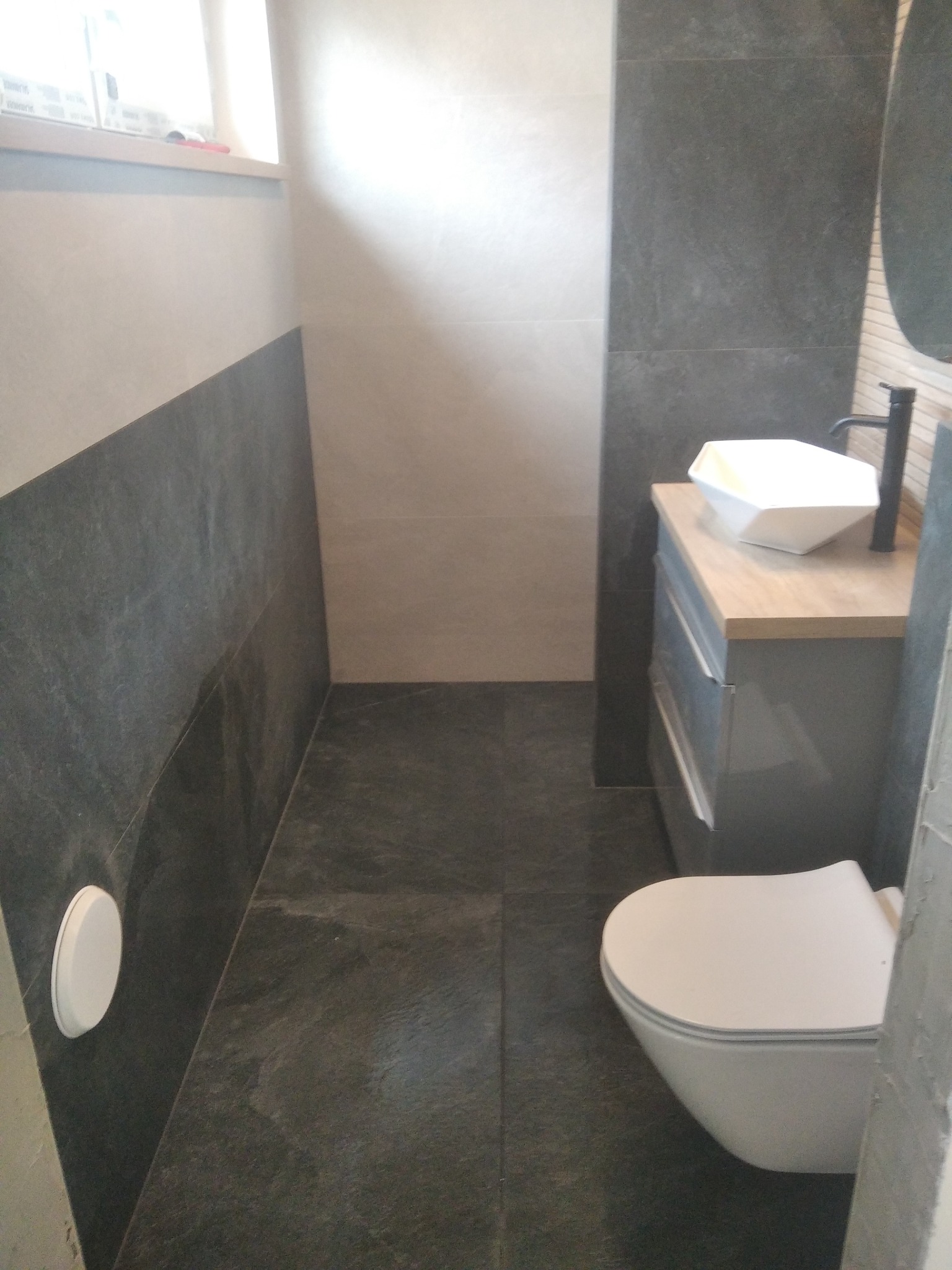 Wykończona łazienka w szaro-białej kolorystyce z umywalką nablatową o geometrycznym kształcie, nowoczesną toaletą i płytkami imitującymi kamień na ścianach i podłodze.