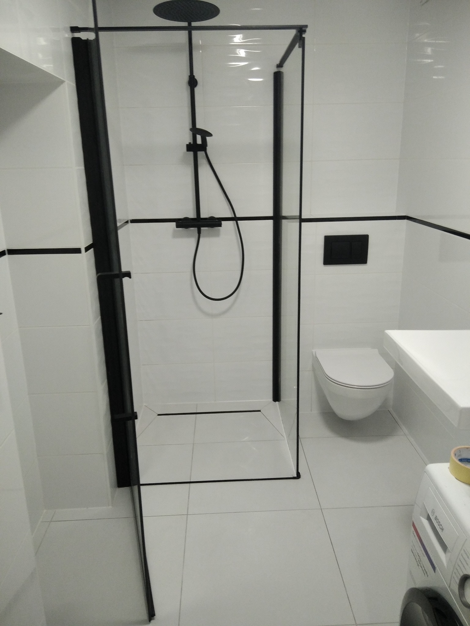 Nowoczesna łazienka z kabiną prysznicową typu walk-in z czarnymi profilami, białe płytki i armatura. Minimalistyczny design z pralką w rogu.