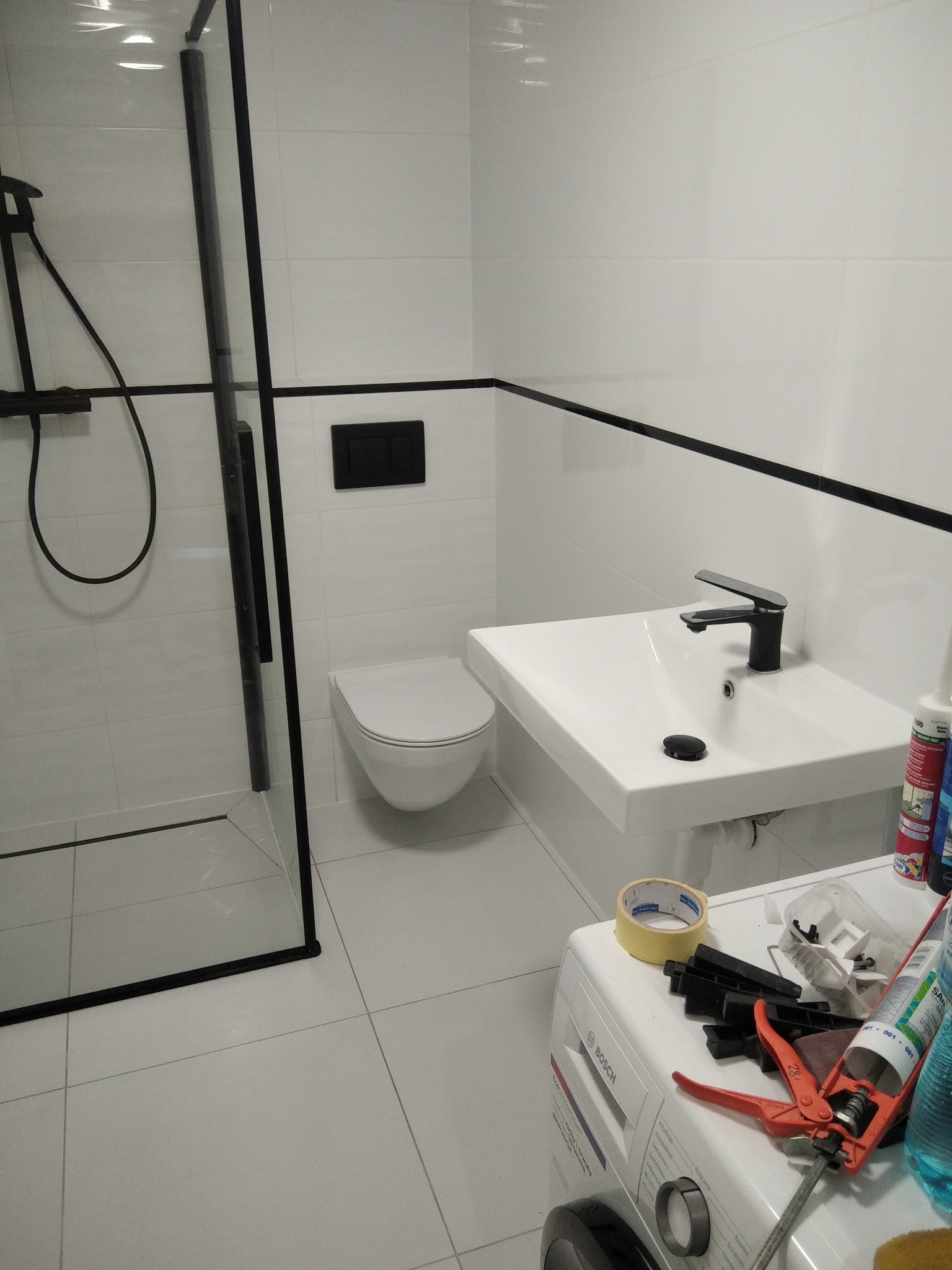 Nowoczesna łazienka z czarnymi akcentami: kabina prysznicowa, umywalka i toaleta. Na pralce narzędzia glazurnicze, podkreślające etap prac wykończeniowych.