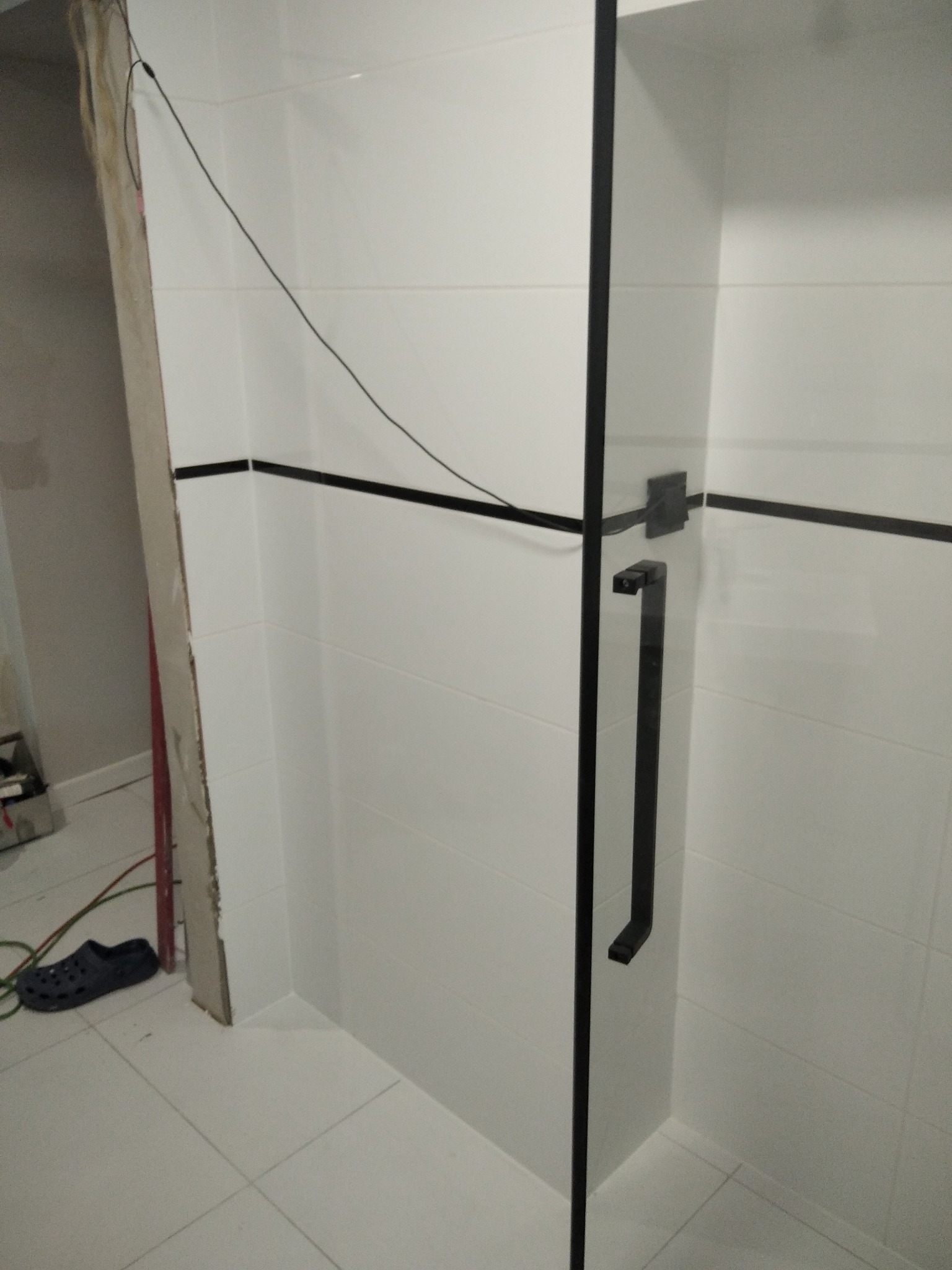 Białe płytki z czarnymi akcentami w wyremontowanej łazience. Widoczna czarna rama kabiny prysznicowej i fragment ściany w trakcie wykończenia. Na podłodze leżą robocze buty.