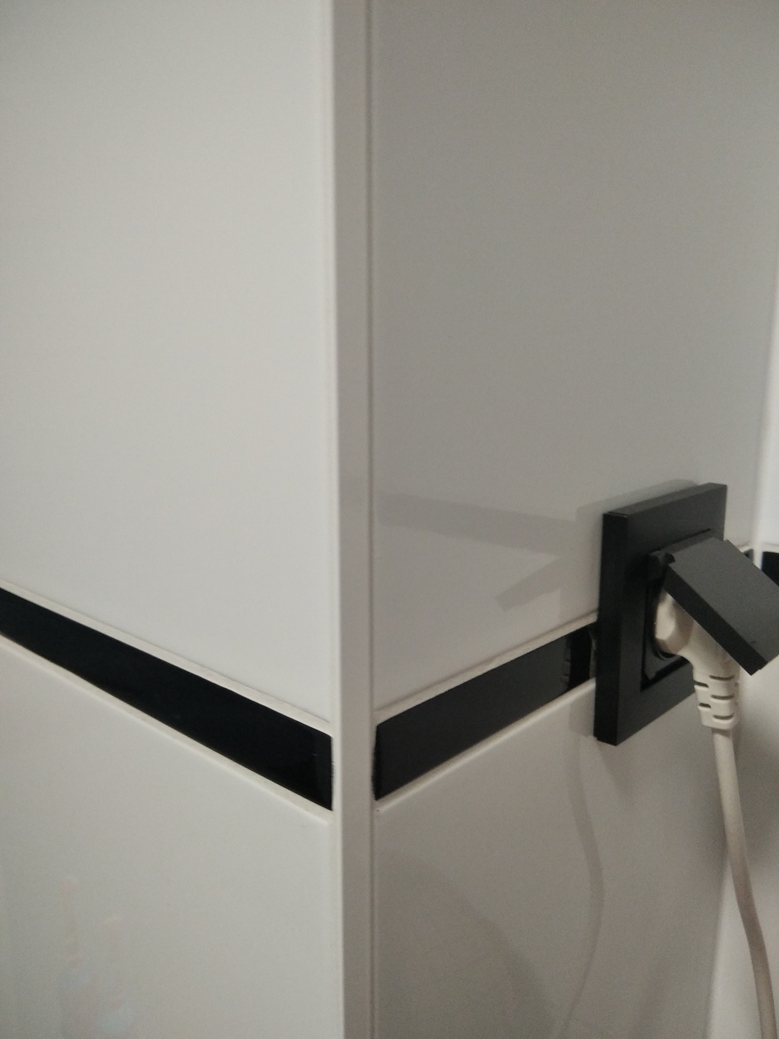 Białe, błyszczące płytki z czarnym pasem dekoracyjnym, narożnik i czarne gniazdko z wtyczką. Minimalistyczne wykończenie ściany w łazience lub kuchni.