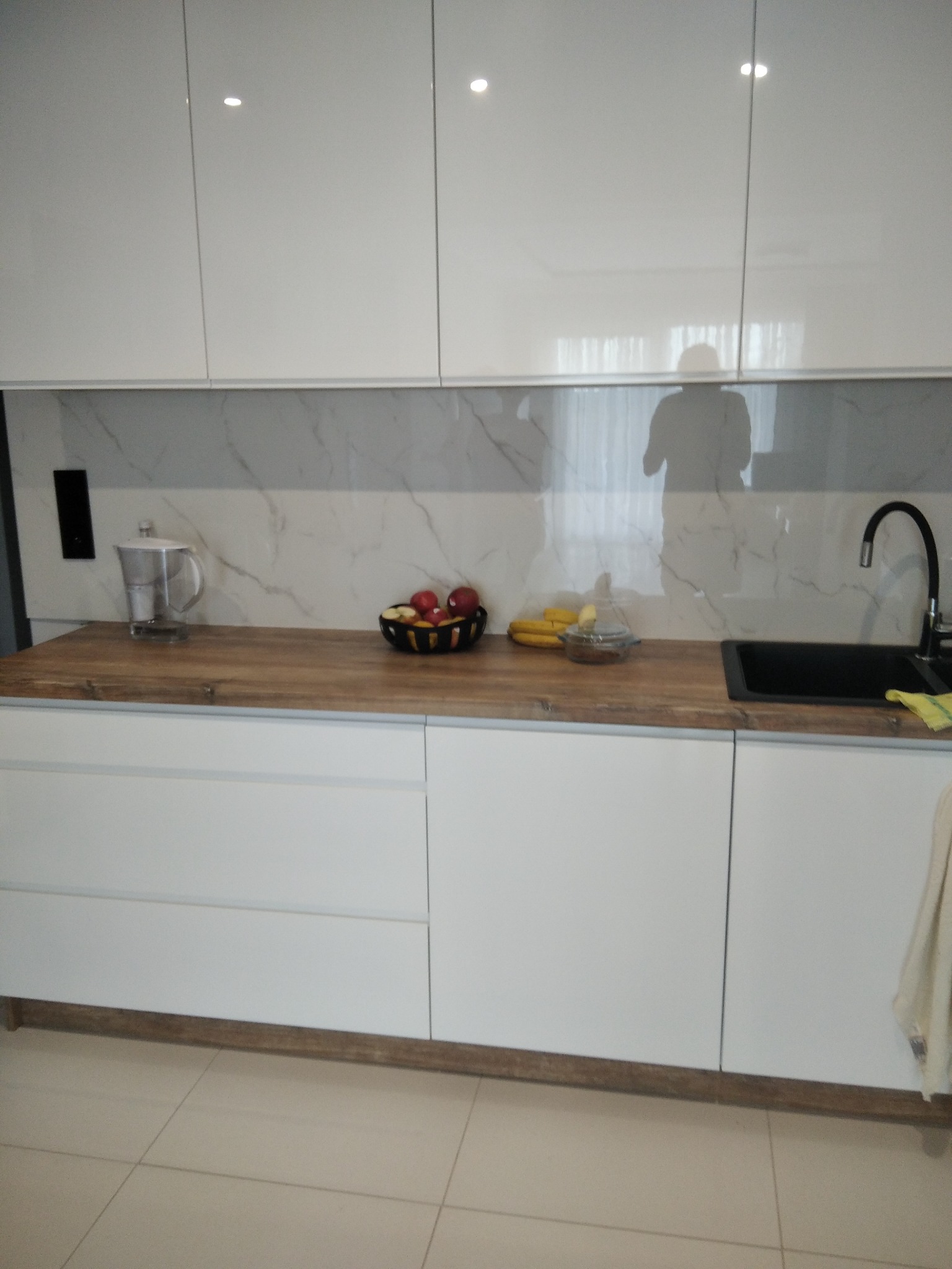 Biała, nowoczesna kuchnia z drewnianym blatem i marmurowym panelem. Na blacie owoce, czajnik i czarna bateria. Widoczny refleks osoby w górnych szafkach.