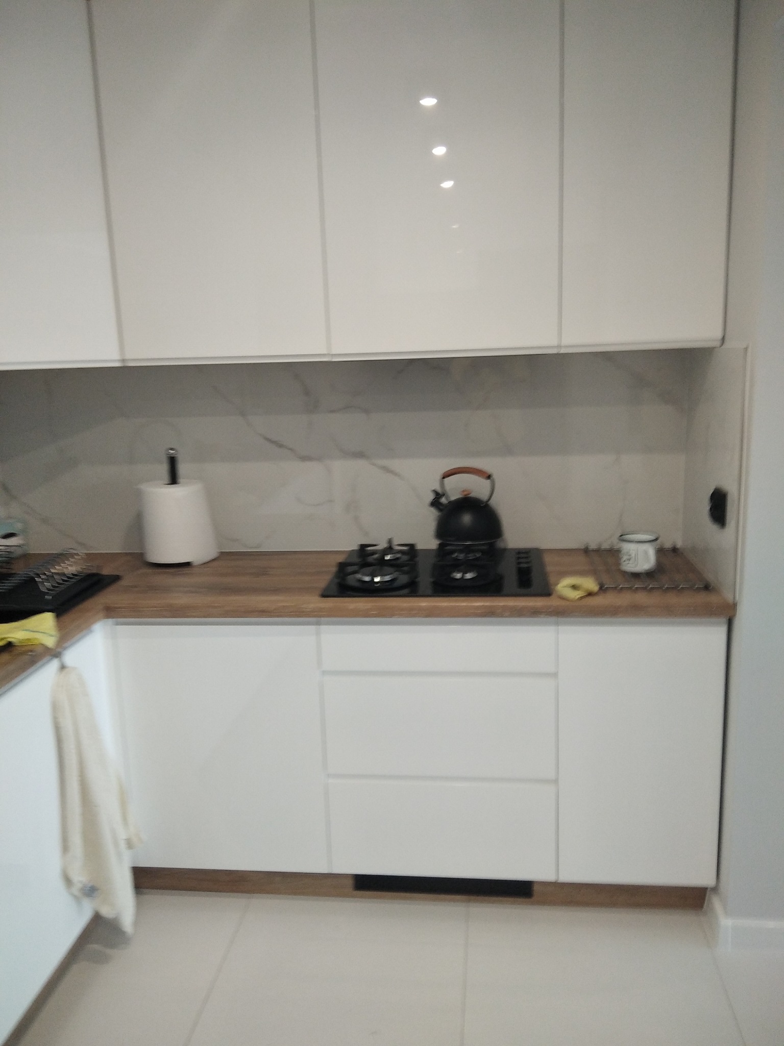 Biała, nowoczesna kuchnia z drewnianym blatem i marmurowym backsplash. Czajnik na płycie gazowej, minimalistyczny design, jasne oświetlenie, widok z perspektywy.