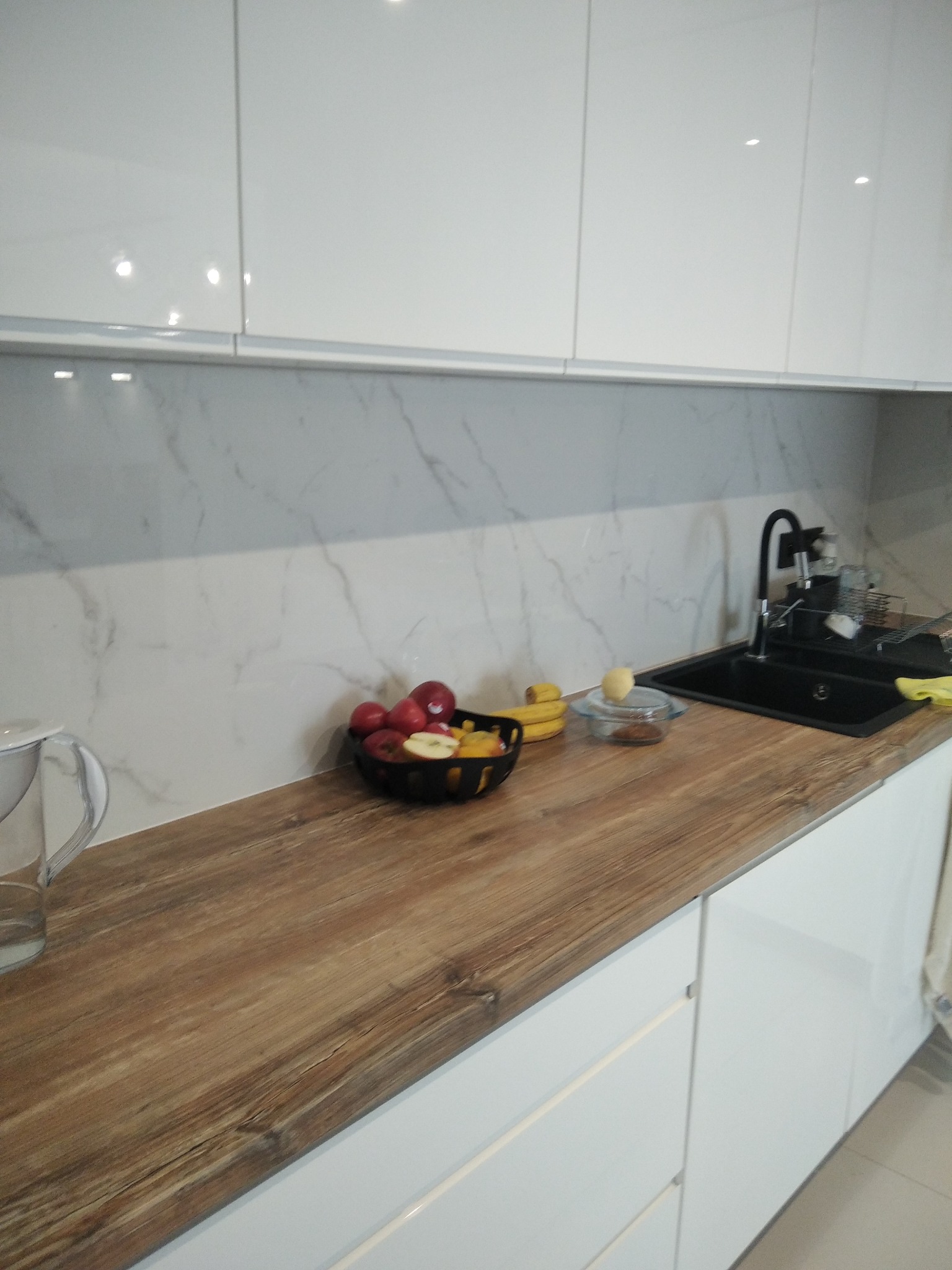 Nowoczesna kuchnia z białymi szafkami i drewnianym blatem. Na blacie owoce w misce, podkreślające domowy charakter. Marmurowe wykończenie ściany nad blatem.