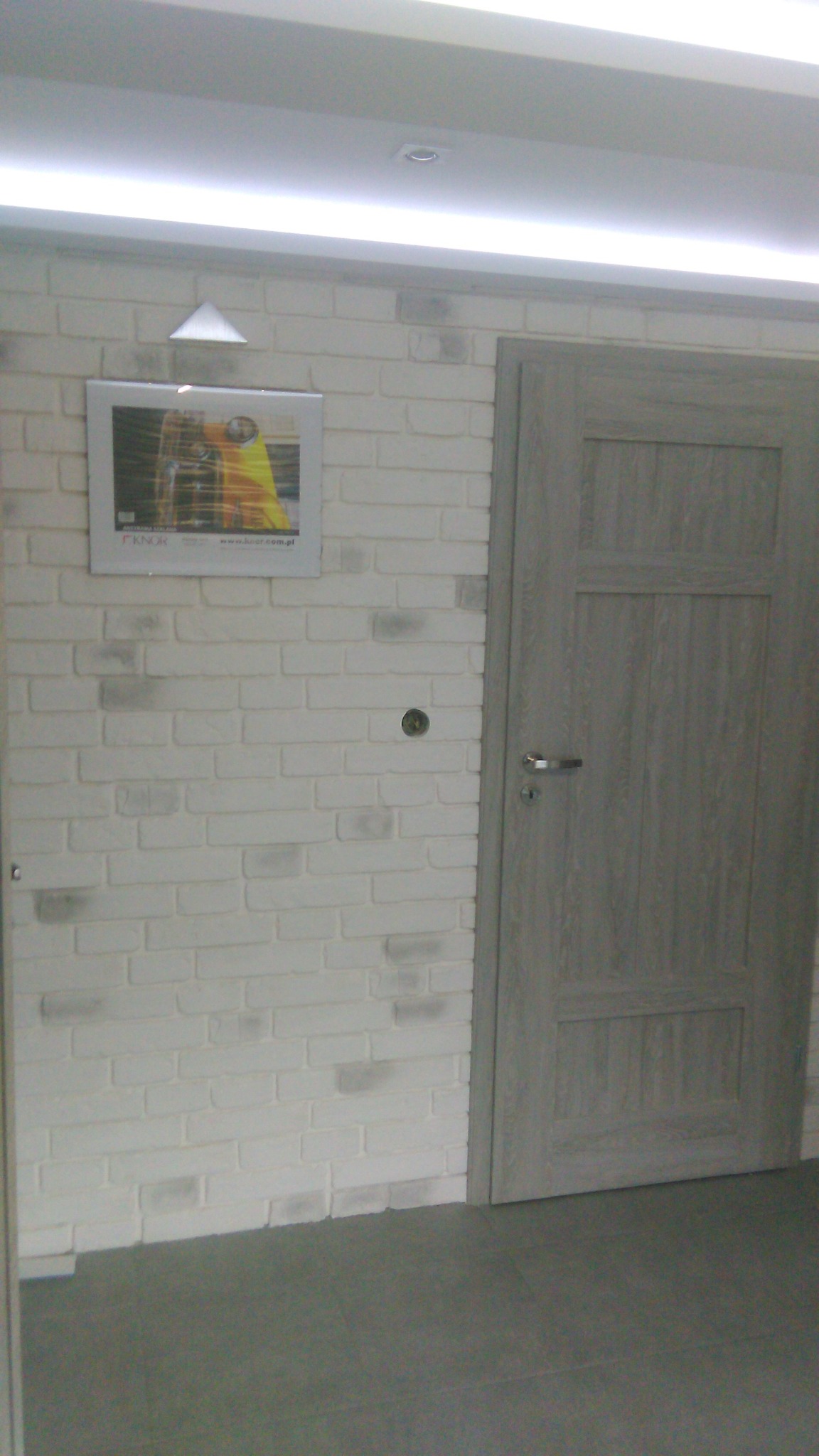 Wnętrze: biała ściana z cegły dekoracyjnej, szara, drewniana płycina drzwiowa z metalową klamką, szara podłoga. Nad ścianą widoczny fragment sufitu z oświetleniem.