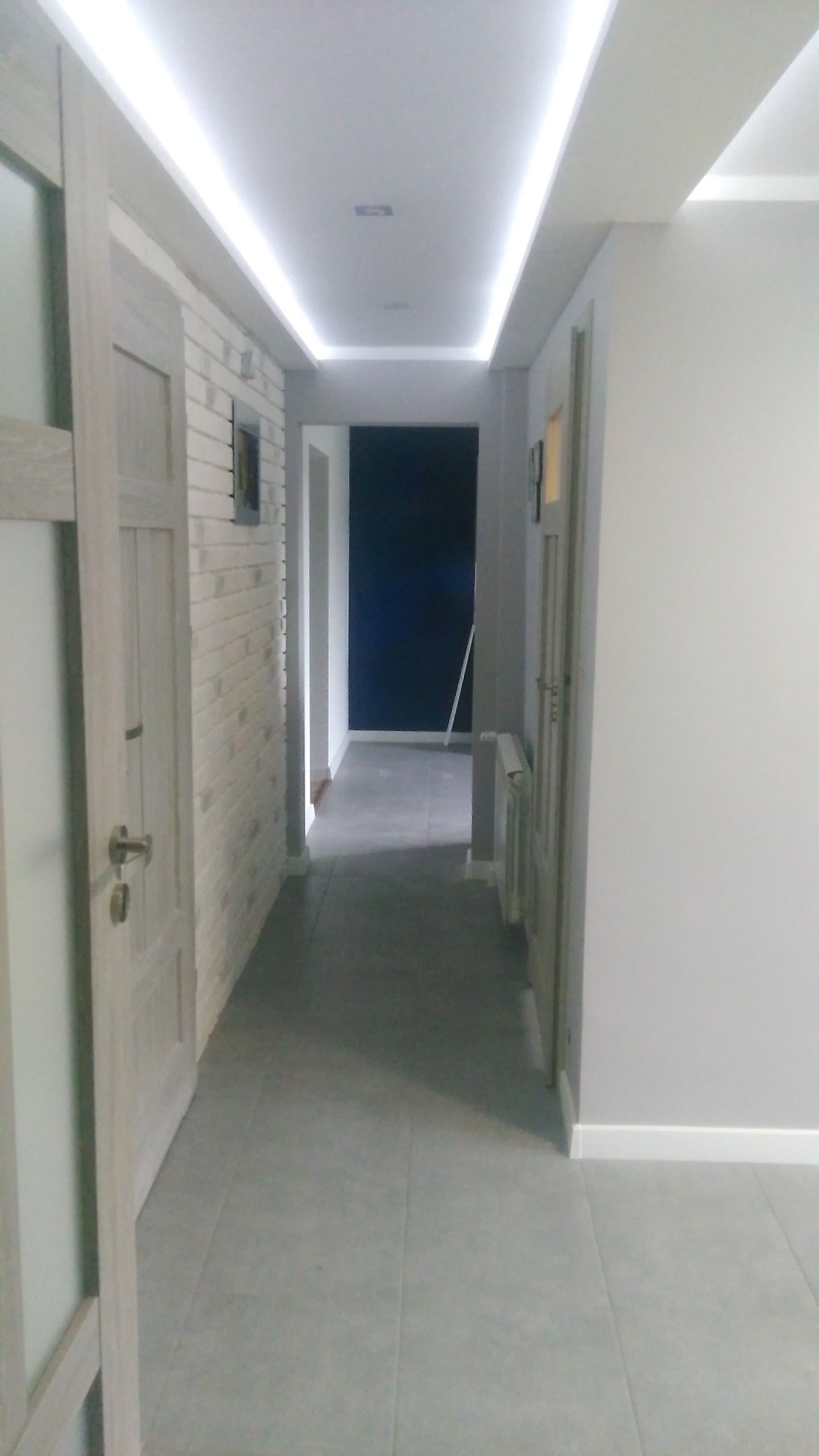 Wykończony korytarz w odcieniach szarości z oświetleniem LED na suficie i imitacją cegły na ścianie. Widoczne drzwi i płytki podłogowe.