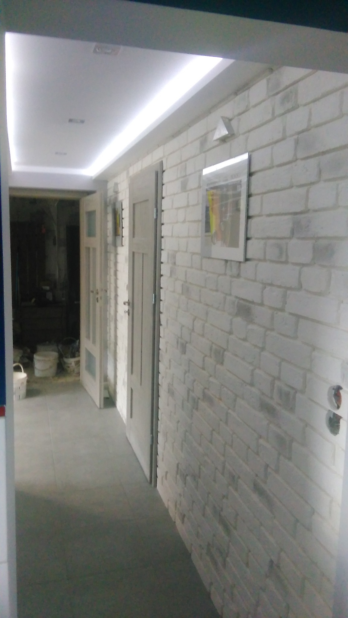 Korytarz po remoncie: biała cegła na ścianie, szare płytki na podłodze, dwoje drzwi wewnętrznych i oświetlenie sufitowe LED. Widoczne puszki elektryczne.