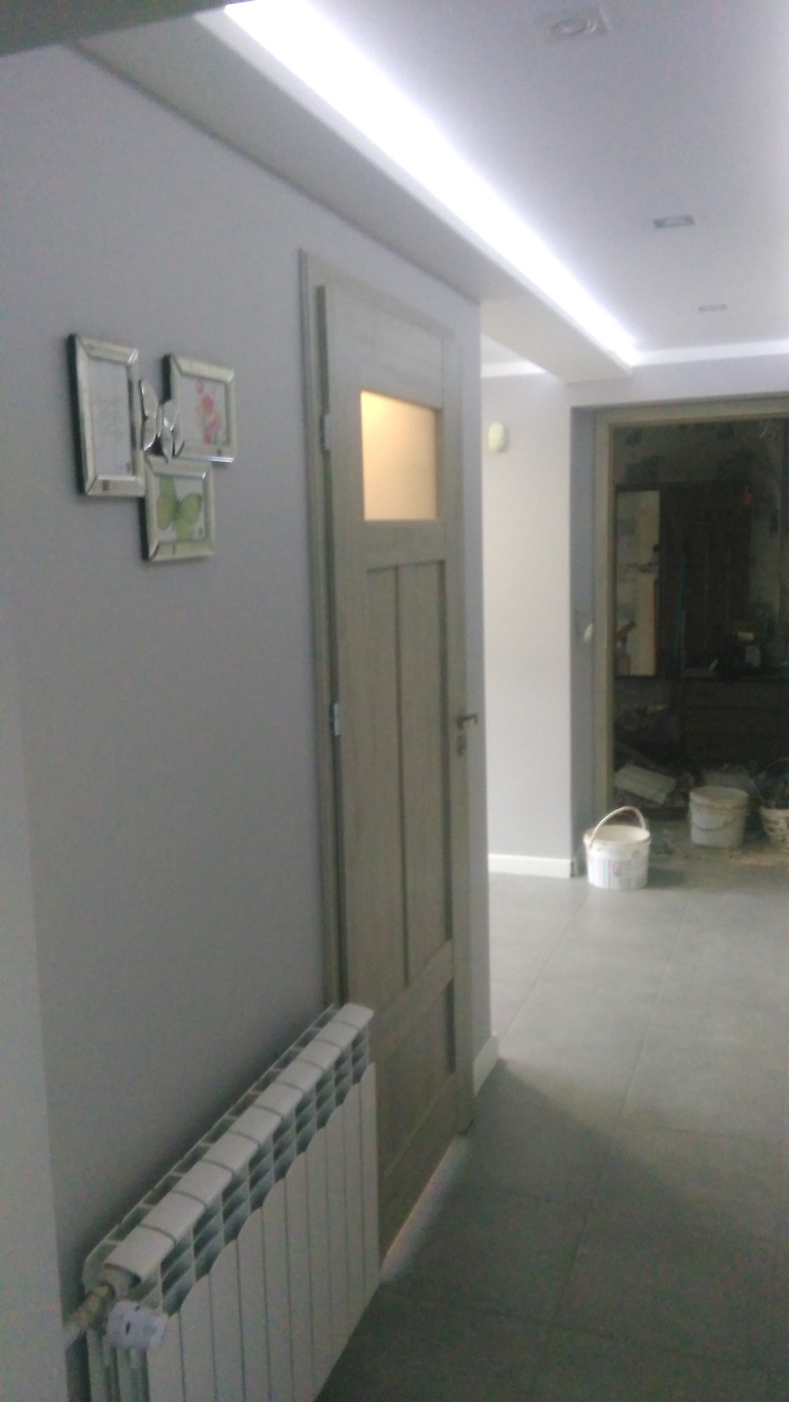 Szara ściana z ramkami, drewniane drzwi z szybką i grzejnik w Grodzisku Wielkopolskim. Widok na wnętrze mieszkania po remoncie. Oświetlenie sufitowe.