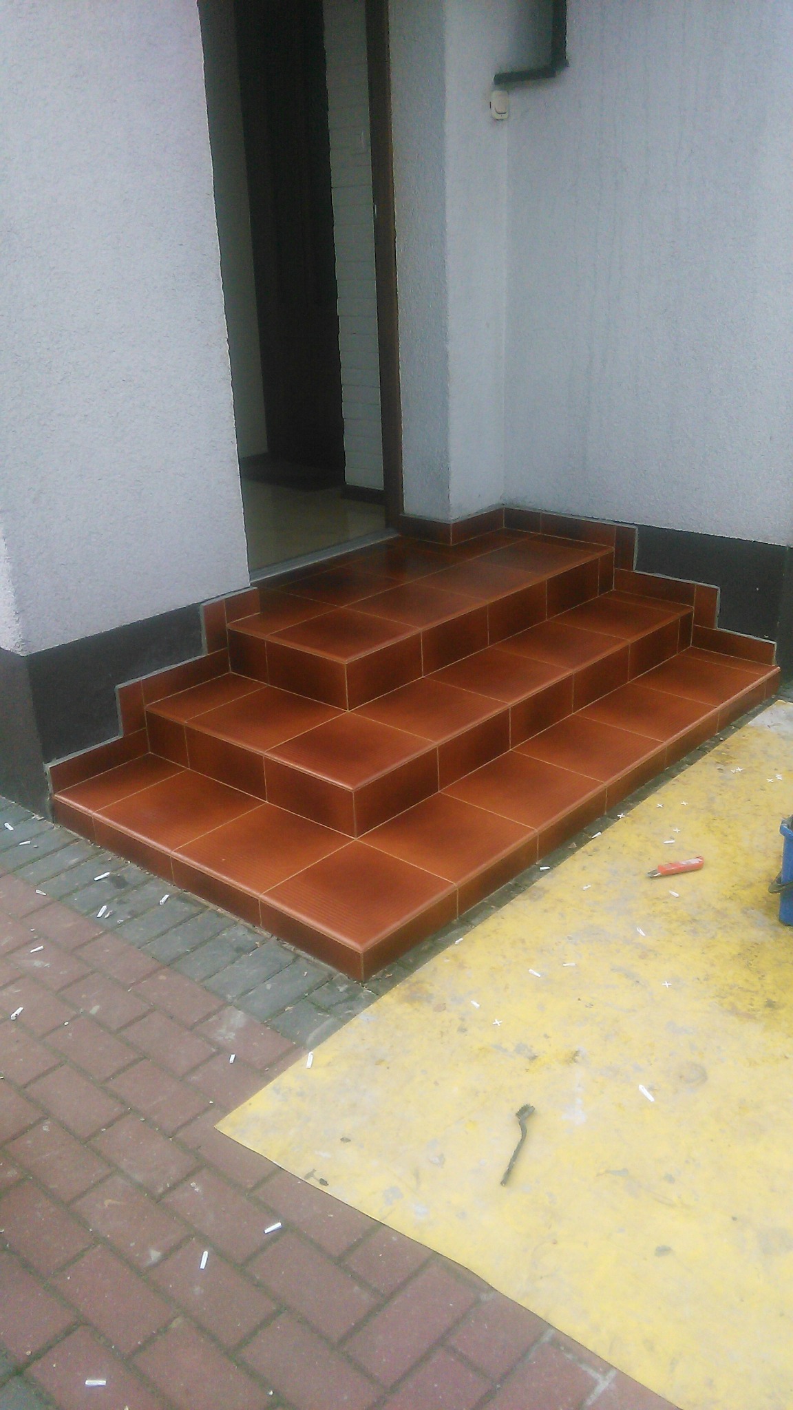 Wykończone, brązowe płytki na schodach wejściowych do budynku. Widoczne fugi, narożniki i fragment chodnika z kostki. Na podłożu narzędzia glazurnicze.