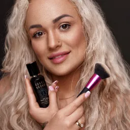 Portret kobiety z blond włosami, trzymającej podkład Bobbi Brown i pędzel do makijażu na ciemnym tle. Widoczne tatuaże na ramionach i złota biżuteria.