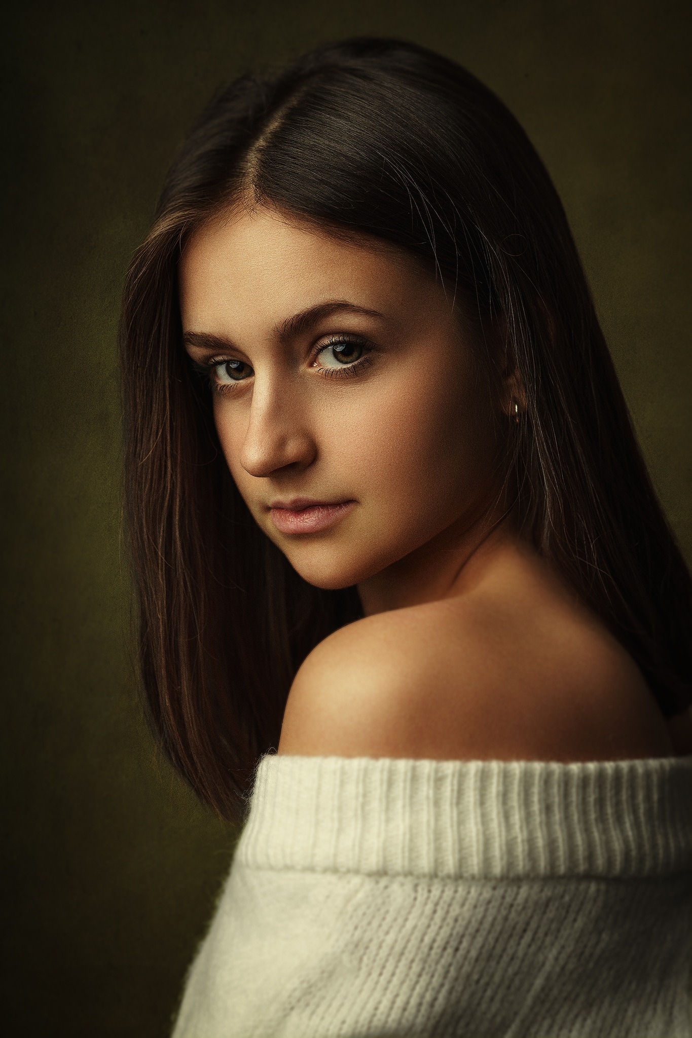 Portret młodej kobiety z długimi, brązowymi włosami, spoglądającej przez ramię, ubrana w biały sweter z odkrytymi ramionami na ciemnym, rozmytym tle.