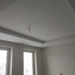 Biały sufit podwieszany z dekoracyjnym obramowaniem w nowym, jasnym pomieszczeniu z dwoma oknami, widoczne przewody elektryczne.