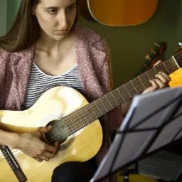 Młoda kobieta w swetrze gra na gitarze klasycznej, patrząc na nuty na stojaku w ciepłym oświetleniu pokoju, z widocznymi innymi instrumentami w tle.