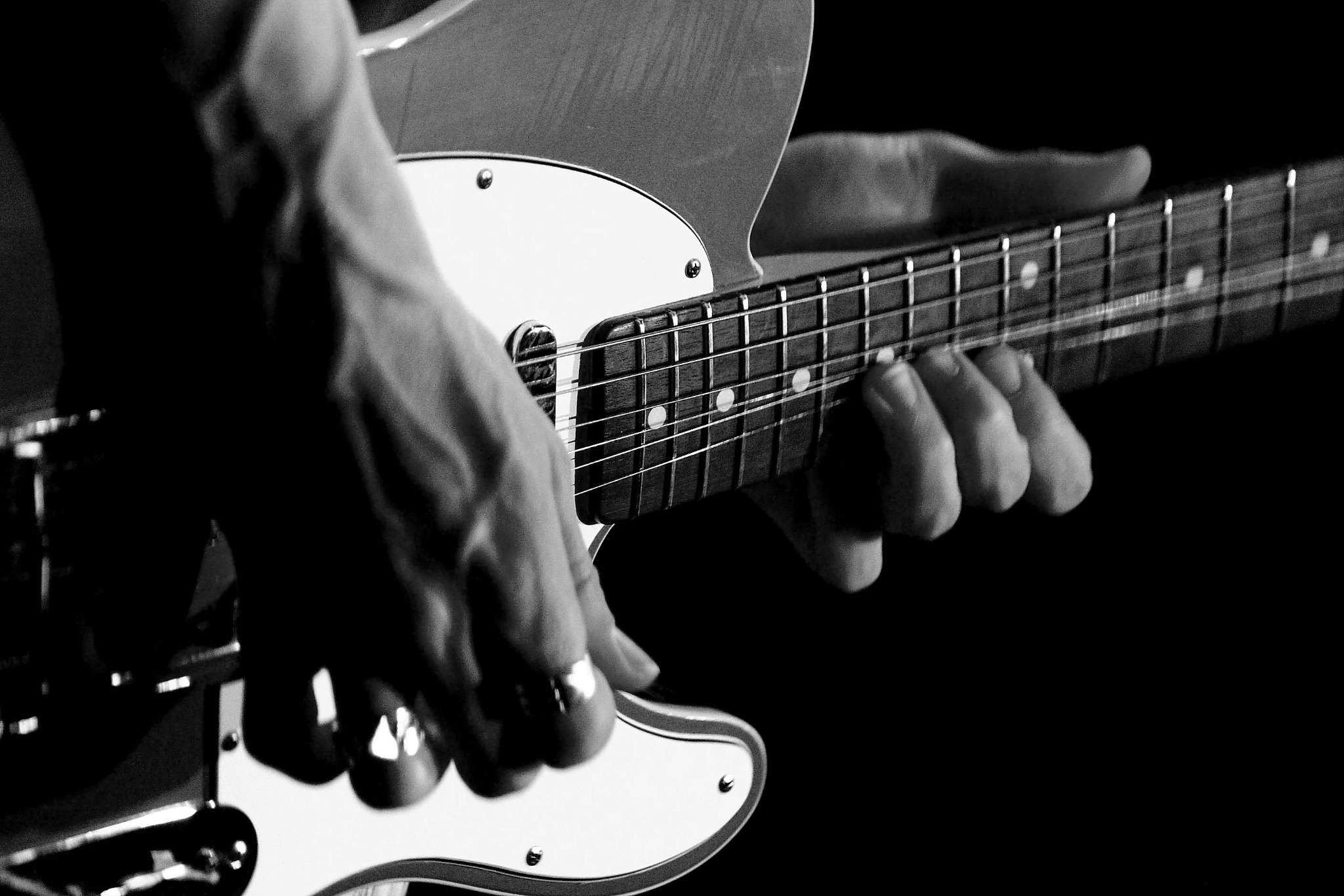 Czarno-białe zbliżenie dłoni grających na elektrycznej gitarze Telecaster, z pierścieniem na palcu wskazującym lewej ręki i ciemnym tłem.