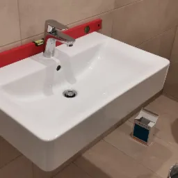 Biała, prostokątna umywalka zamontowana na ścianie z użyciem poziomicy dla precyzyjnego ustawienia, widoczna bateria i syfon, obok toaleta w folii ochronnej i karton po ceramice, na ścianie beżowe...