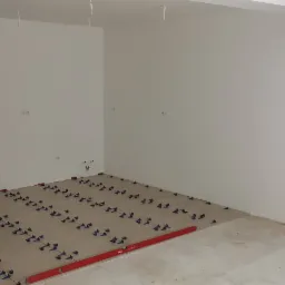 Przygotowanie do układania płytek podłogowych w pustym, białym pomieszczeniu; widoczny laserowy poziom, krzyżyki dystansowe i fragment ułożonej płyty. Pracownik w roboczym ubraniu stoi przy wejściu.