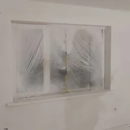 Świeżo pomalowane na biało wnętrze pokoju z zabezpieczonym folią oknem i widocznymi śladami gładzi na ścianach.